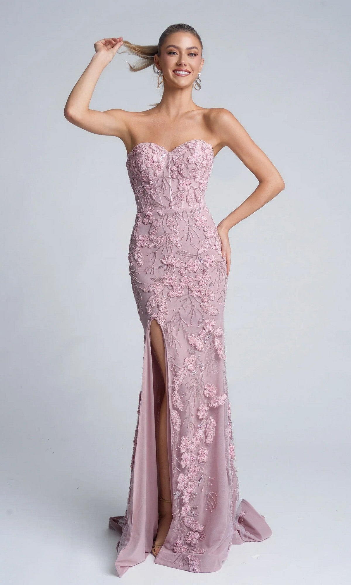 A woman stands smiling in the pink Portia and Scarlett PS26074 long formal dress with a strapless sweetheart neckline, floral embroidery, and a thigh-high slit, holding her ponytail with one hand.